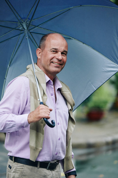 Szczęśliwy dojrzały mężczyzna chodzenie w The ulica z duży parasol pod The deszcz - Zdjęcie, obraz