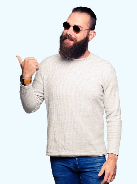 Young hipster man wearing sunglasses smiling with happy face looking and pointing to the side with thumb up. - Photo, Image