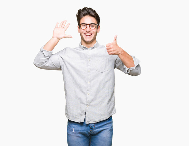 Jonge knappe man met bril geïsoleerd achtergrond weergegeven en met de vingers omhoog nummer zes terwijl lacht zelfverzekerd en blij. - Foto, afbeelding