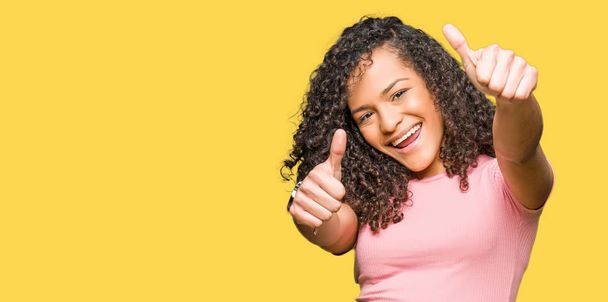Young beautiful woman with curly hair wearing pink t-shirt approving doing positive gesture with hand, thumbs up smiling and happy for success. Looking at the camera, winner gesture. - Photo, Image