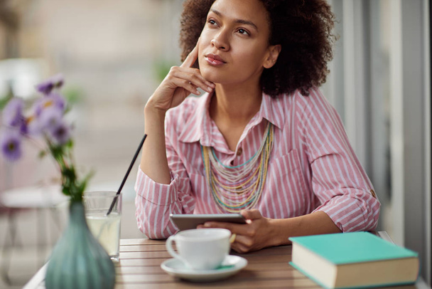 Grogeous mixed race nainen vaaleanpunainen raidallinen mekko istuu kahvilassa ja käyttää tabletti. Pöydällä on kuppi kahvia, kirjaa ja limonadia
. - Valokuva, kuva