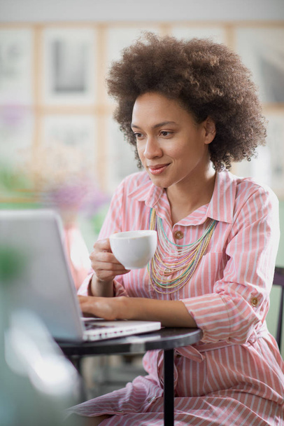 Bájos vegyes verseny nő csíkos rózsaszín ruhában aitting a kávézóban, kávét inni és használata laptop. - Fotó, kép