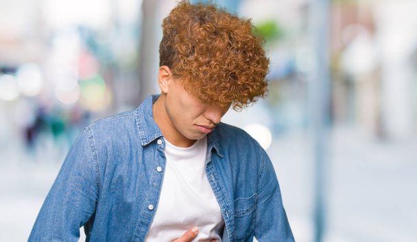 Young handsome man with afro hair wearing denim jacket with hand on stomach because nausea, painful disease feeling unwell. Ache concept. - Photo, Image