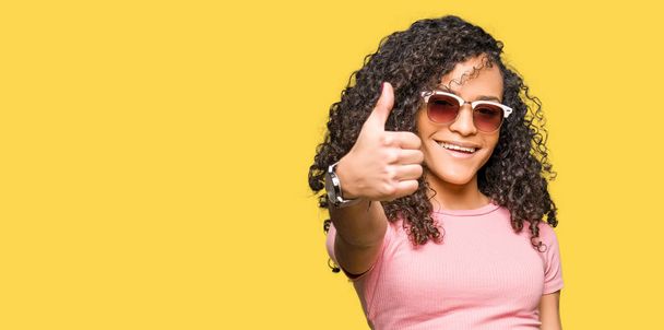 Young beautiful woman with curly hair wearing pink sunglasses doing happy thumbs up gesture with hand. Approving expression looking at the camera with showing success. - Fotografie, Obrázek