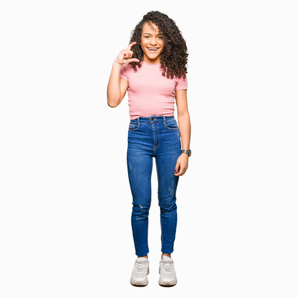 Young beautiful woman with curly hair wearing pink t-shirt smiling and confident gesturing with hand doing size sign with fingers while looking and the camera. Measure concept. - Фото, изображение