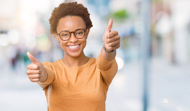 Młode piękne african american Kobieta w okularach na na białym tle zatwierdzające robi pozytywny gest ręką, kciuki, uśmiechając się i szczęśliwy dla sukcesu. Patrząc na kamery, zwycięzca gest. - Zdjęcie, obraz