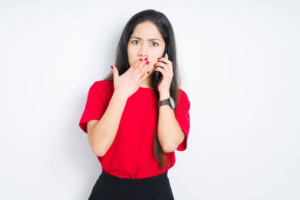 Νεαρή μελαχρινή γυναίκα που μιλάει στο smartphone πάνω από το απομονωμένο στόμα κάλυμμα του στόματος με το χέρι σοκαρισμένος από την ντροπή για το λάθος, έκφραση του φόβου, φοβισμένος στη σιωπή, μυστική έννοια - Φωτογραφία, εικόνα