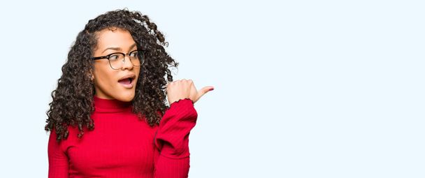 Young beautiful woman with curly hair wearing glasses smiling with happy face looking and pointing to the side with thumb up. - Fotografie, Obrázek