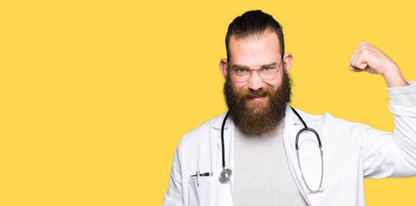 Jovem médico loiro com barba vestindo casaco médico Pessoa forte mostrando músculo do braço, confiante e orgulhoso do poder
 - Foto, Imagem