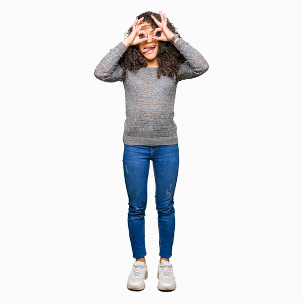 Young beautiful woman with curly hair wearing grey sweater doing ok gesture like binoculars sticking tongue out, eyes looking through fingers. Crazy expression. - Foto, afbeelding