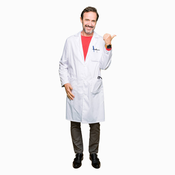 Therapeutin mittleren Alters im weißen Mantel lächelt mit glücklichem Gesicht und zeigt mit erhobenem Daumen zur Seite. - Foto, Bild