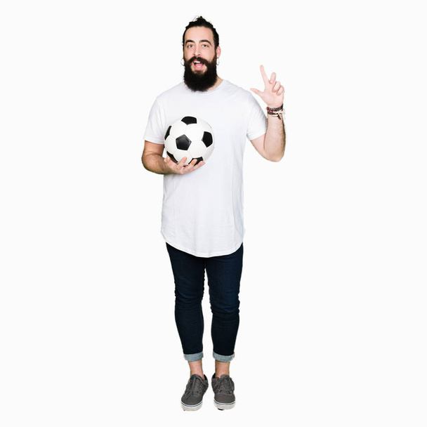 Young man with long hair and bear holding soccer football ball surprised with an idea or question pointing finger with happy face, number one - Zdjęcie, obraz