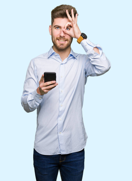 Όμορφος νεαρός επαγγελματική χρήση smartphone με χαρούμενο πρόσωπο χαμογελά κάνει εντάξει σημάδι με το χέρι στο μάτι που αναζητούν μέσα από τα δάχτυλα - Φωτογραφία, εικόνα