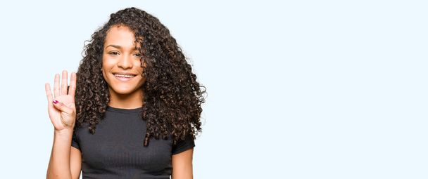 Young beautiful woman with curly hair showing and pointing up with fingers number four while smiling confident and happy. - Photo, Image