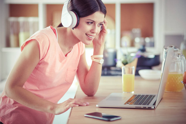 Портрет веселої молодої жінки, яка слухає музику з навушниками і використовує ноутбук, стоячи на кухні
 - Фото, зображення