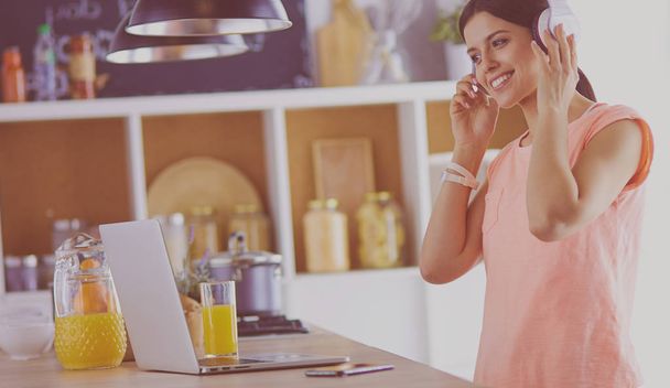 Портрет веселой молодой женщины, слушающей музыку в наушниках и пользующейся ноутбуком, стоя на кухне
 - Фото, изображение