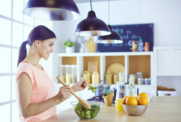 Smiling young woman mixing fresh salad in the kitchen. - Foto, imagen
