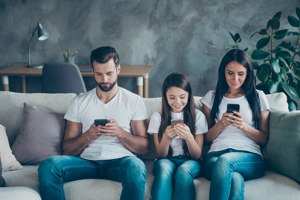 Portrait of nice attractive lovely cheerful charming focused family wearing casual white t-shirts jeans denim sitting on sofa using gadget device at industrial style interior living-room - Foto, immagini
