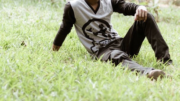 Zavřít oříznutý obraz mladého muže v příležitostným oblečení, které sedí v parku na zelené trávě, v ranním denní době. Pohled zepředu. Koncept trávení volného času v otevřeném vzduchu obklopeném přírodou. H - Fotografie, Obrázek