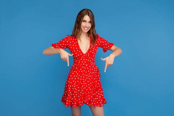 Pretty young woman with bright smile in red dress pointing fingers down over blue background. - Foto, Imagem