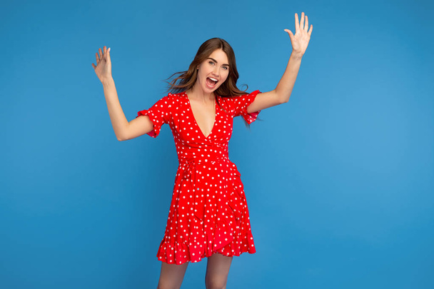 Crazy young woman with bright smile in red casual dress looking at camera with excited face with hands up over blue background. - Foto, Imagen