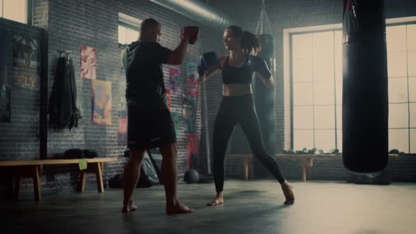 Fit Athletic Woman Kickboxer Punches and Hits the Punching Pads During a Workout in a Loft Gym. She's Beautiful and Energetic. Strong Coach is Holding the Boxing Pads. Intense Self-Defence Training. - Πλάνα, βίντεο