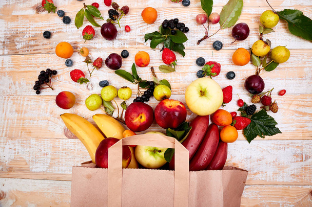 Paper bag of different health fruits food on wooden background. Top view. Flat lay Beige canvas grocery bag fallen over while dropping fruits - Photo, Image