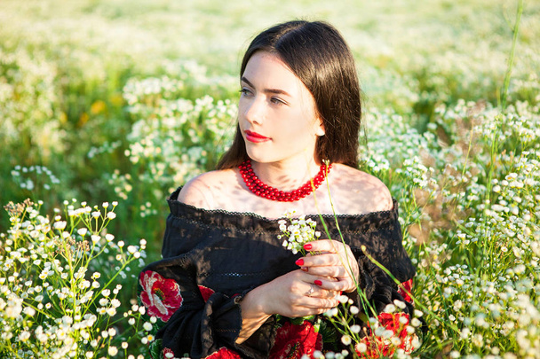 Εμφάνιση ενός κοριτσιού που κάθεται σε ένα χωράφι με λουλούδια, ιεροτελεστία του πέρασμα, εμφάνιση ενός νεαρού κοριτσιού στην ουκρανική στολή, κόκκινες χάντρες, κορίτσι στο πεδίο, κορίτσι κοιτάζει ένα μπουκέτο, εισπνέετε το άρωμα των λουλουδιών, νεαρό κορίτσι βρίσκεται στο πεδίο, Κοίτα ένα νεαρό κορίτσι Sniffing, - Φωτογραφία, εικόνα