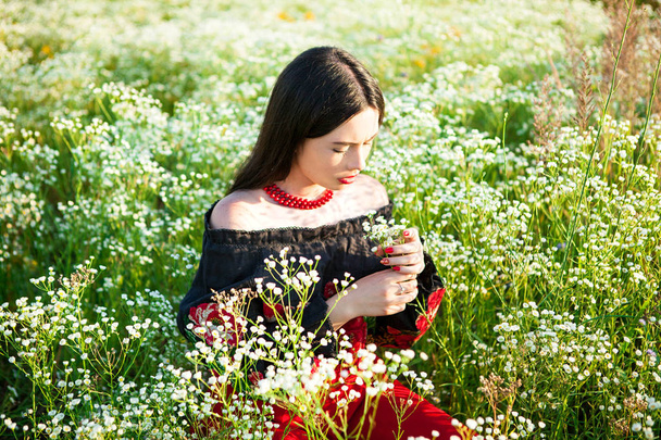 Mädchen sieht einen kleinen Strauß Wildblumen auf dem Feld, Mädchen schaut auf einen Strauß, inhaliert den Duft der Blumen, riecht die Blumen, junges Mädchen liegt auf dem Feld, Blick eines jungen Mädchens, bestickte Bluse, Blick eines jungen Mädchens im ukrainischen Outfit,  - Foto, Bild