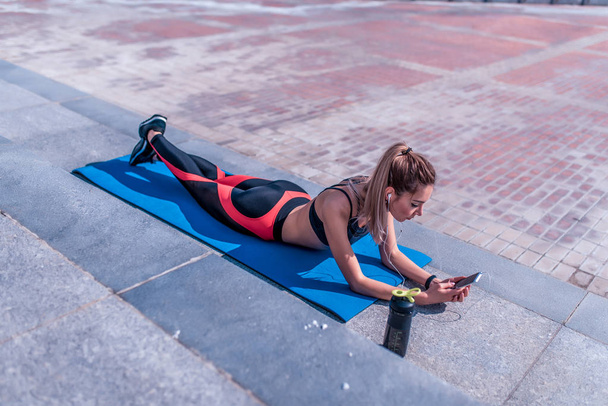 sports girl summer city, lies on yoga mat, listens watches online training, video podcast, writes message on phone, online app on Internet, leisure fitness jogging workouts morning. Sportswear. - Fotó, kép