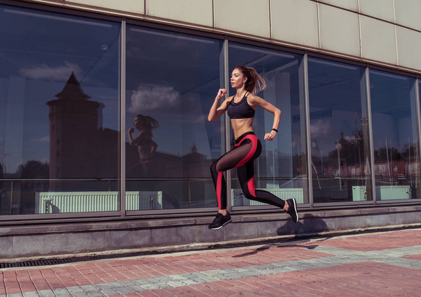 Tanned woman, fitness instructor is jumping girl athlete is jogging in morning, in summer city, beautiful athlete is engaged in fitness, youth lifestyle, healthy in city. Free space for text. - Photo, Image