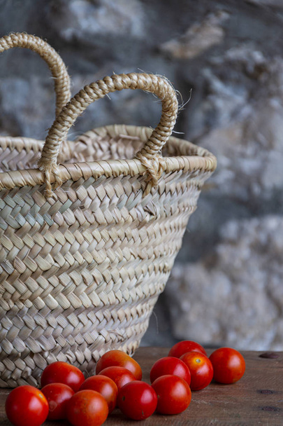 Close-up of cherry tomatoes on wooden table, straw basket and stone background in vertical - Photo, Image
