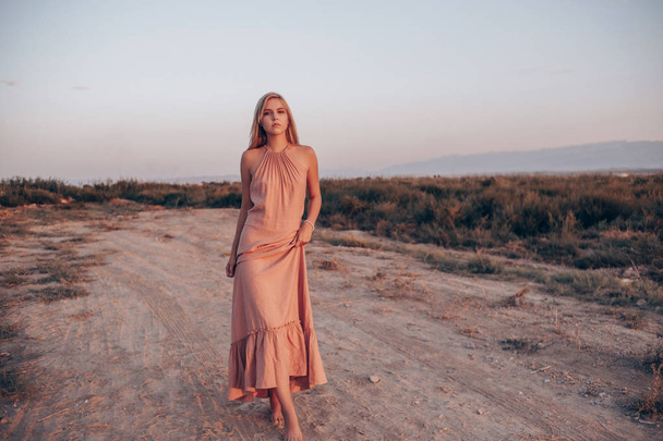nuori kaunis valkoihoinen nainen vaaleanpunainen mekko kävelee hiekalla
 - Valokuva, kuva