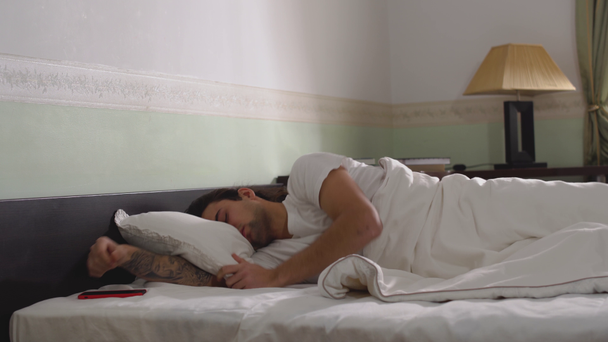 Man asleep in the wide bed wakes up looks at the phone and goes to bed and falls asleep again but then quickly jumps out of bed. - Imágenes, Vídeo