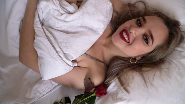 βίντεο με νεαρή χαμογελαστή κυρία με φωτεινό μακιγιάζ ξαπλωμένη στο κρεβάτι από κοντά. Θηλυκά μάτια κοιτάζουν την κάμερα. Το κόκκινο τριαντάφυλλο απλώνεται κοντά  - Πλάνα, βίντεο