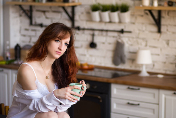 Adorable nice charming cheerful pretty trendy stylish girl drinking coffee, sitting on table countertop in modern light white kitchen - Foto, afbeelding