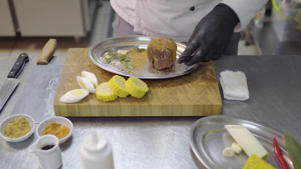 Шеф-повара в черных латексных перчатках втирают кусок телятины в металлическую пластину, маринуя мясо. Кухонный стол с посудой и ингредиентами
. - Кадры, видео