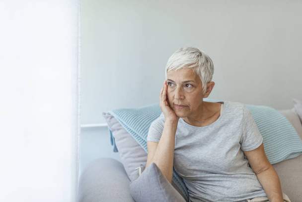 Sad Mature Woman Suffering From Agoraphobia Looking Out Of Window. Depressed senior woman sitting on sofa at home. Depressed senior woman at home - Photo, Image