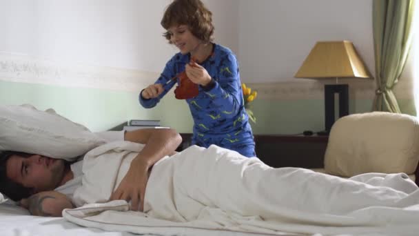 Older brother sleep in bed and small brother wake him up while playing the violin. Sleepy young guy throw pillow at yanger bro and close ears with pillows. - Кадры, видео