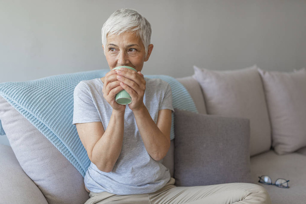 Happy mature woman sitting on couch while drinking cup of coffee. - Foto, immagini