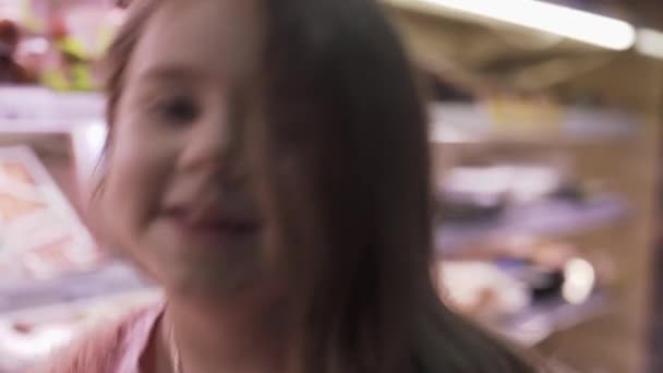 Cheerful little girl shows thumb up on the background of grocery shelves. Little girl in the supermarket. Daughter helps mom shopping in the store. - Filmagem, Vídeo