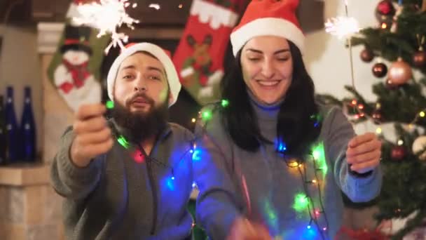 Man and woman in home clothes sitting near new year tree, waving sparklers in hands. Woman gives present box to man, kissing Happy couple celebrates Christmas, video  - Filmagem, Vídeo