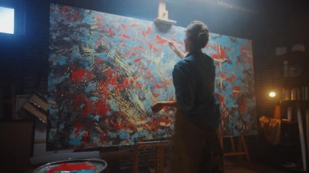 Talented Female Artist Works on Abstract Oil Painting, Using Paint Brush She Creates Modern Masterpiece. Dark and Messy Creative Studio where Large Canvas Stands on Easel Illuminated. Zoom out - Footage, Video