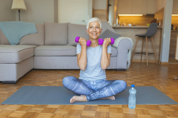 Portrait of senior woman lifting dumbbells. Senior woman doing fitness exercices in front of tv. Happy senior woman making fitness training with dumbbells - Фото, изображение