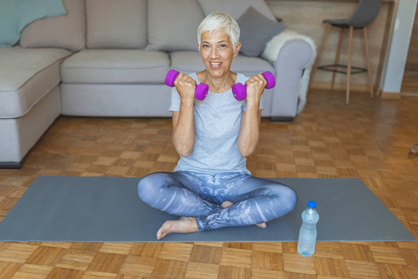 Fit mature woman lifting dumbbell while sitting at home. Mature Woman Exercise At Home. Age is no excuse to slack on your health. Senior Woman Lifting Dumbbells - Fotografie, Obrázek