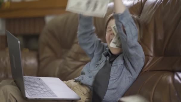 Malý šestiletý chlapec sedí na kožené pohovce s notebookem na nohách a vyhazuje peníze do vzduchu. Práce na internetu. - Záběry, video