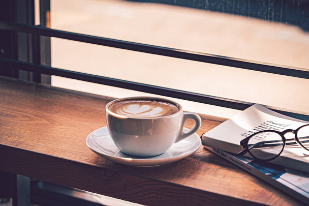 Latte koffie in witte koffie kopje op de houten tafel voor het grote raam van het koffiehuis. - Foto, afbeelding