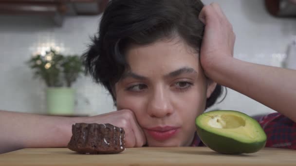 Portrait of hungry beautiful girl choosing between little tasty brownie cupcake with delicious frosting and fresh avocado, video  - Video, Çekim