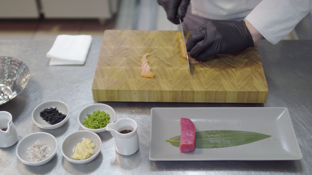 Mãos de chef no uniforme de restaurante branco que corta o pequeno peixe de salmão. Utensílios de cozinha, especiarias, colheres, pedaços de atum, molhos e outros ingredientes
. - Filmagem, Vídeo