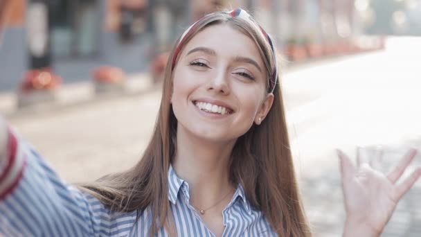 Young Attractive Girl Wearing in Striped Shirt with Headband Waving Hello on Video chat Conversation Selfie with Smartphone and Showing Okey Smiling Standing on The City Street. - Video